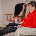 Image shows Kathie who has macular degeneration sitting in an armchair listening to an audiobook and laughing.